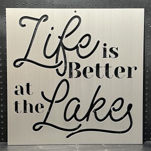 "Life is Better at the Lake" text cutout on brushed metal sign with decorative script against a perforated background.