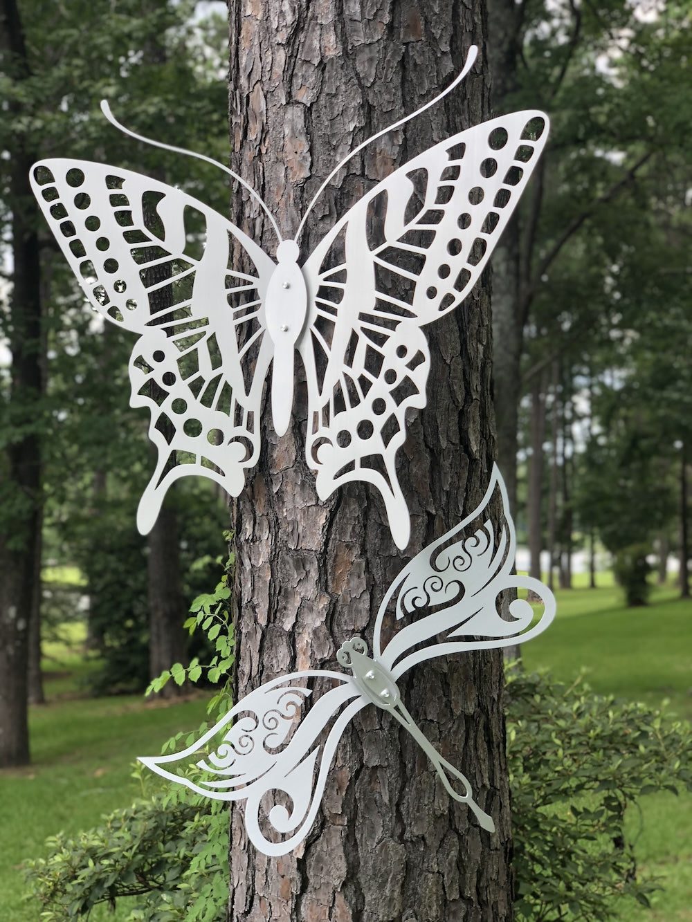 White metal butterfly and dragonfly art with intricate cutouts mounted on a tree, surrounded by a lush forest.