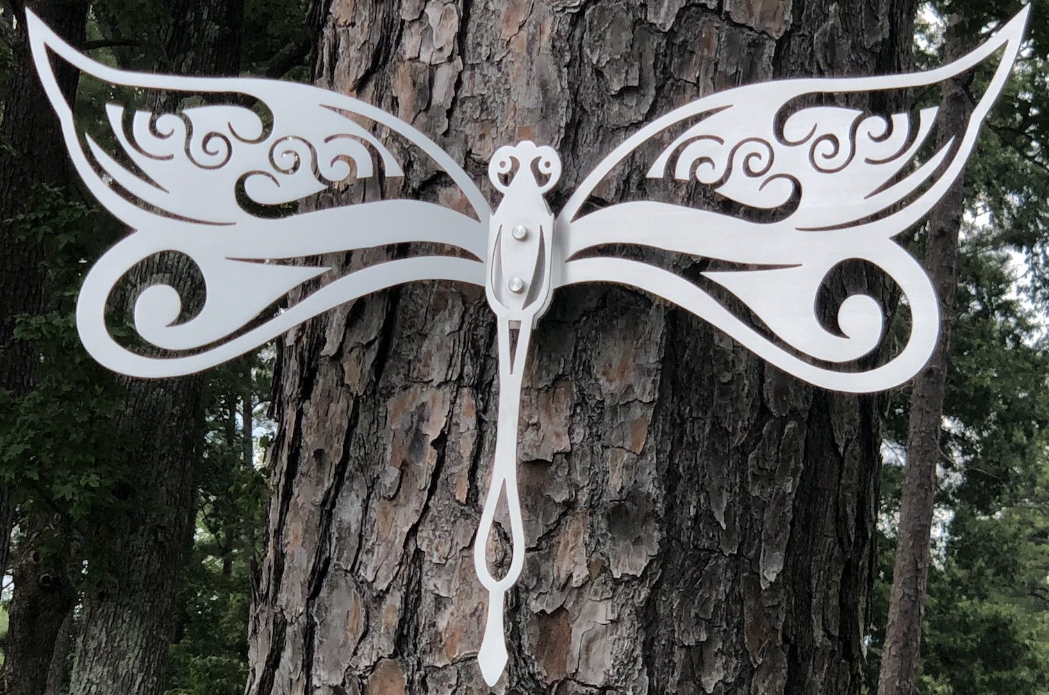White metal dragonfly art on a tree with intricate cutout wings and a forest backdrop.