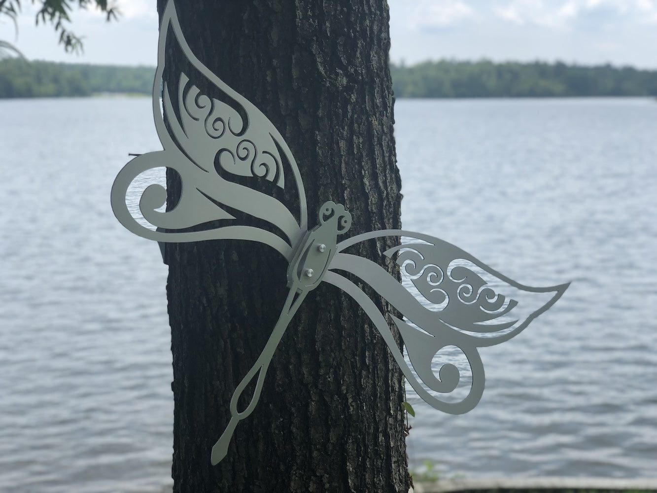 White metal butterfly art with intricate cutouts on a tree by a peaceful lake.