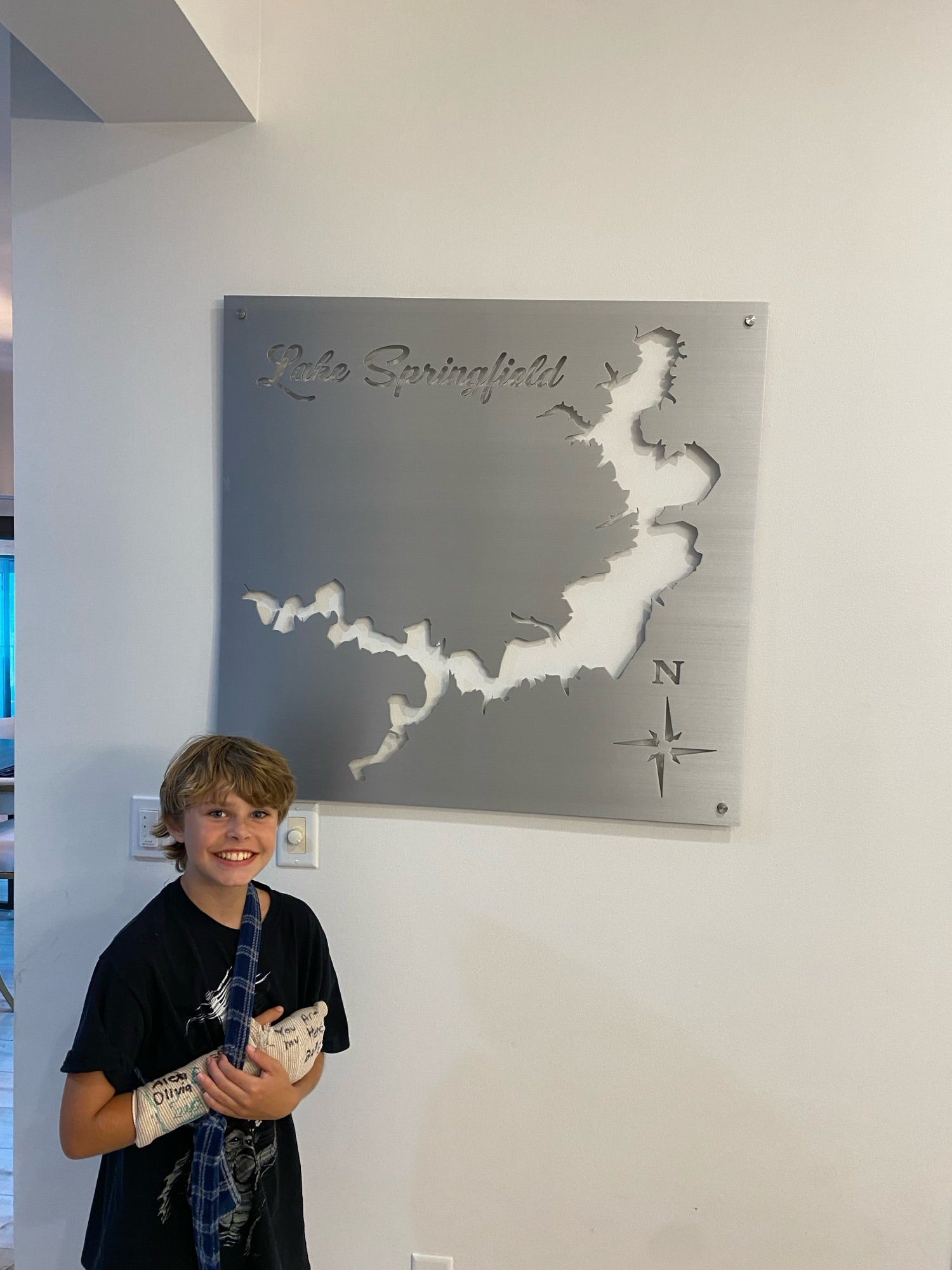 Boy smiling next to a Lake Springfield metal map with compass rose cutout, mounted on a light wall.