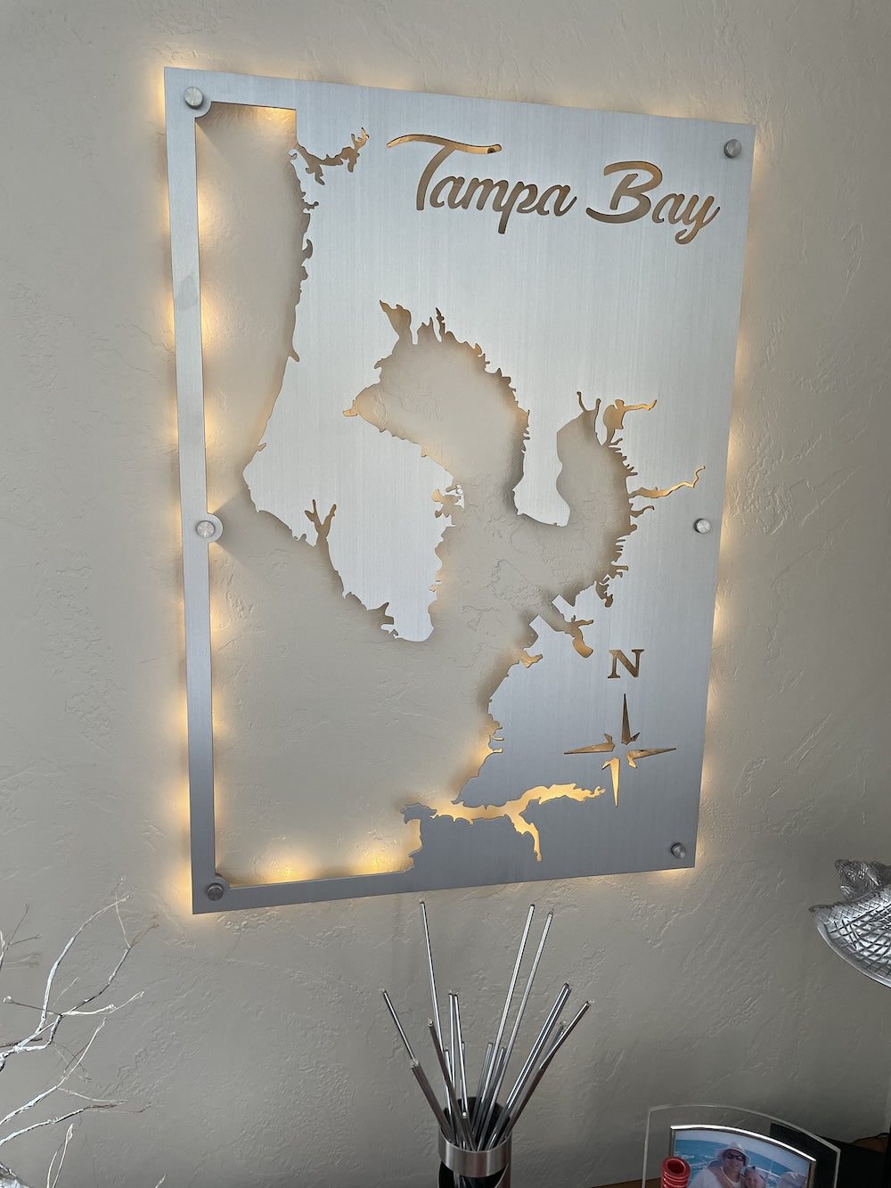Tampa Bay metal map with compass rose, backlit and mounted on a textured wall, above decorative items and a photo.