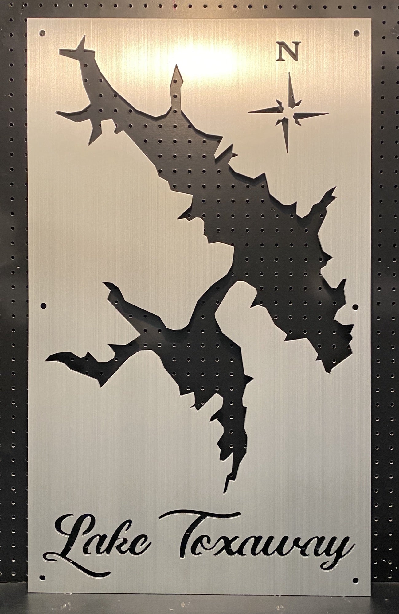 Lake Toxaway custom metal map with compass rose, displayed on a textured black pegboard.