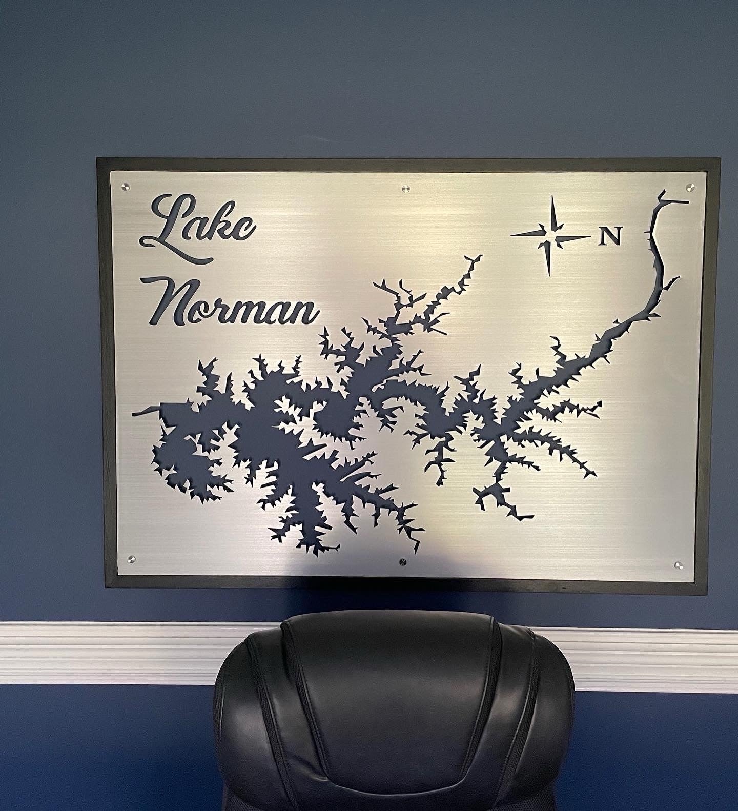 Lake Norman metal map with compass rose on a brushed silver background, mounted on a blue wall above a black chair.
