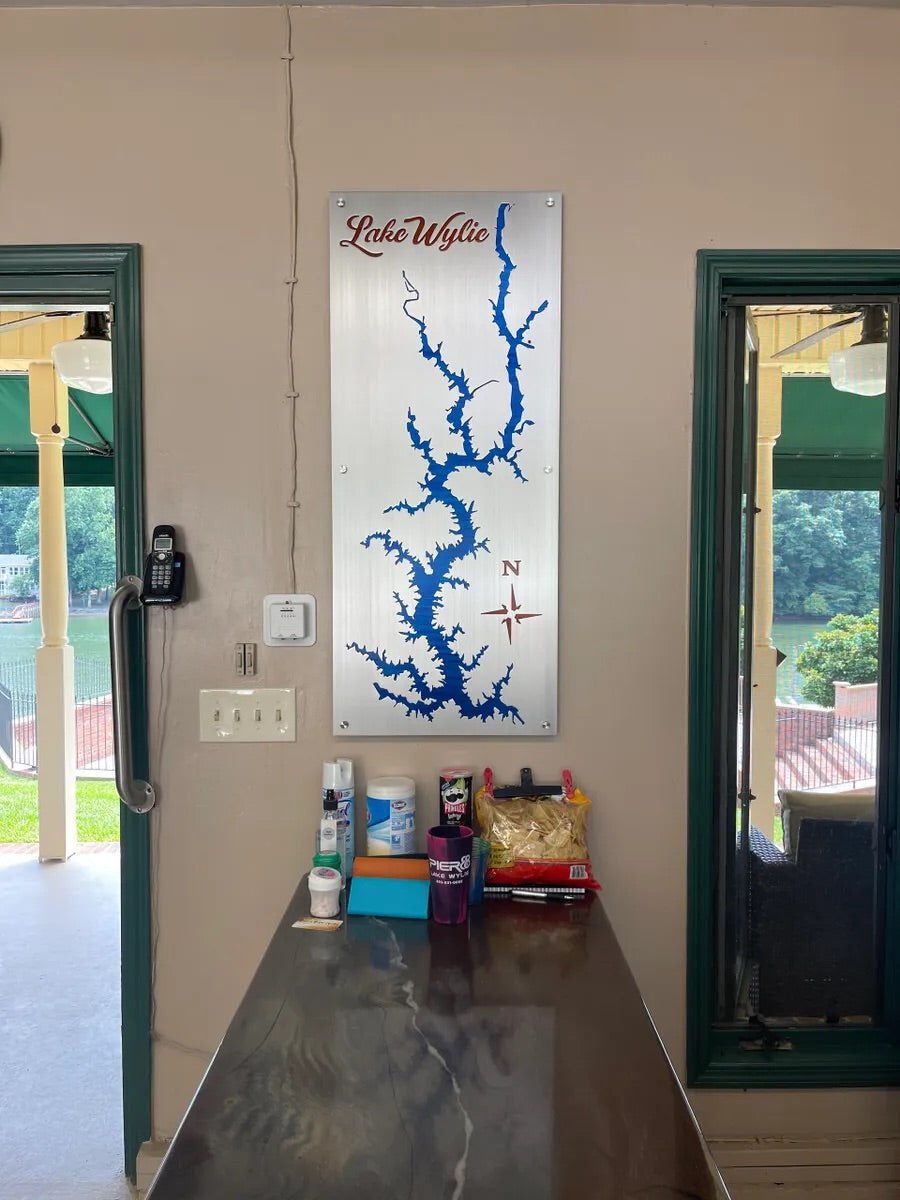 Lake Wylie custom map with blue cutout and compass rose, mounted on a beige wall above a counter with snacks and supplies.