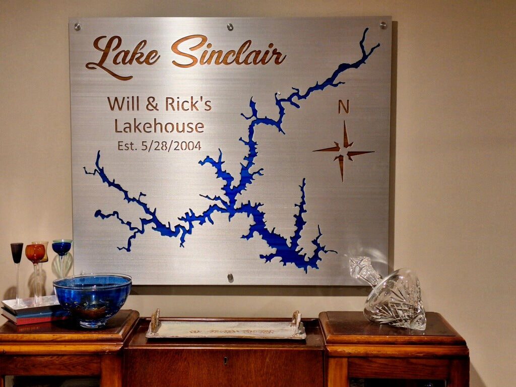 Lake Sinclair metal map with "Will & Rick's Lakehouse Est. 5/28/2004" and compass rose, displayed on a wooden side table.