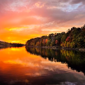 Sunset over a calm lake with colorful fall foliage on trees reflected in the water.