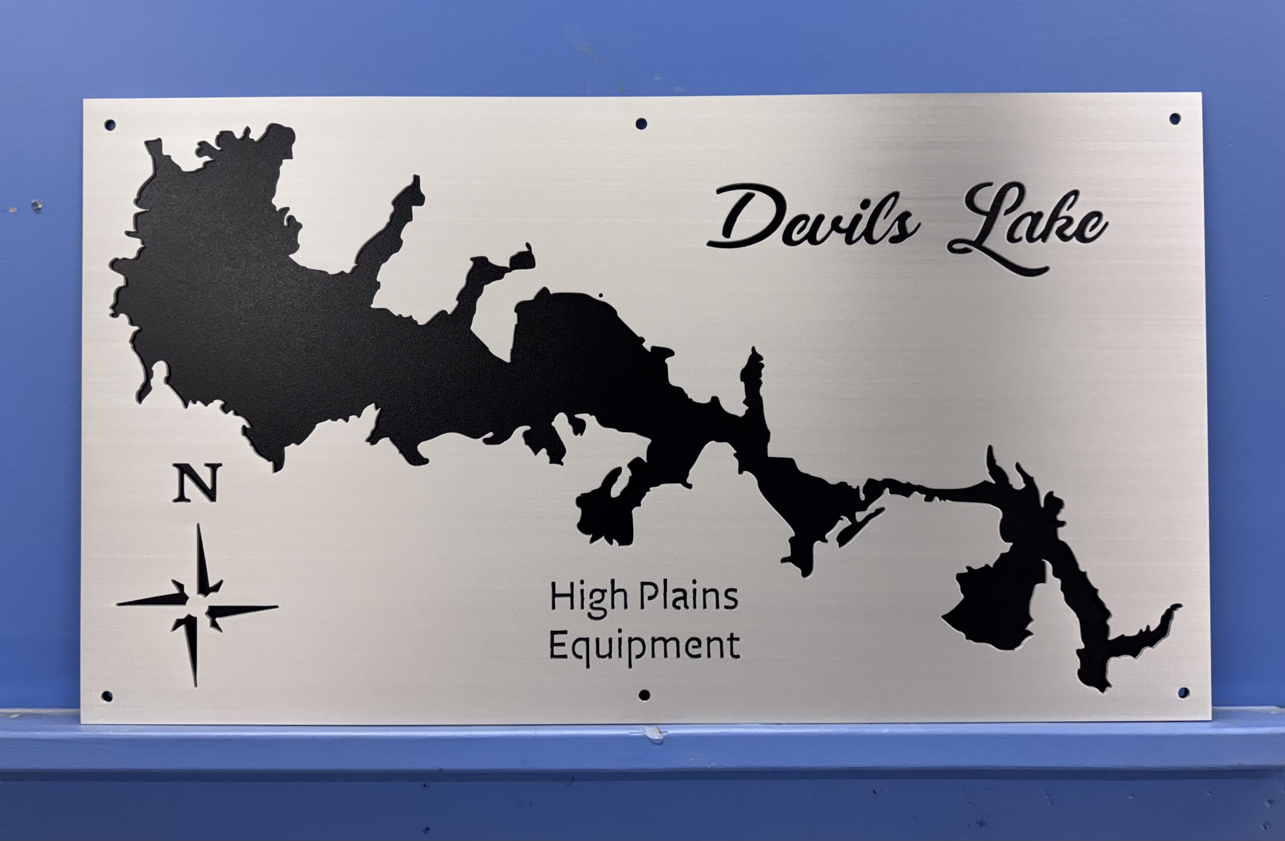 Metal map of Devils Lake with black silhouette and text for High Plains Equipment. North indicator present.