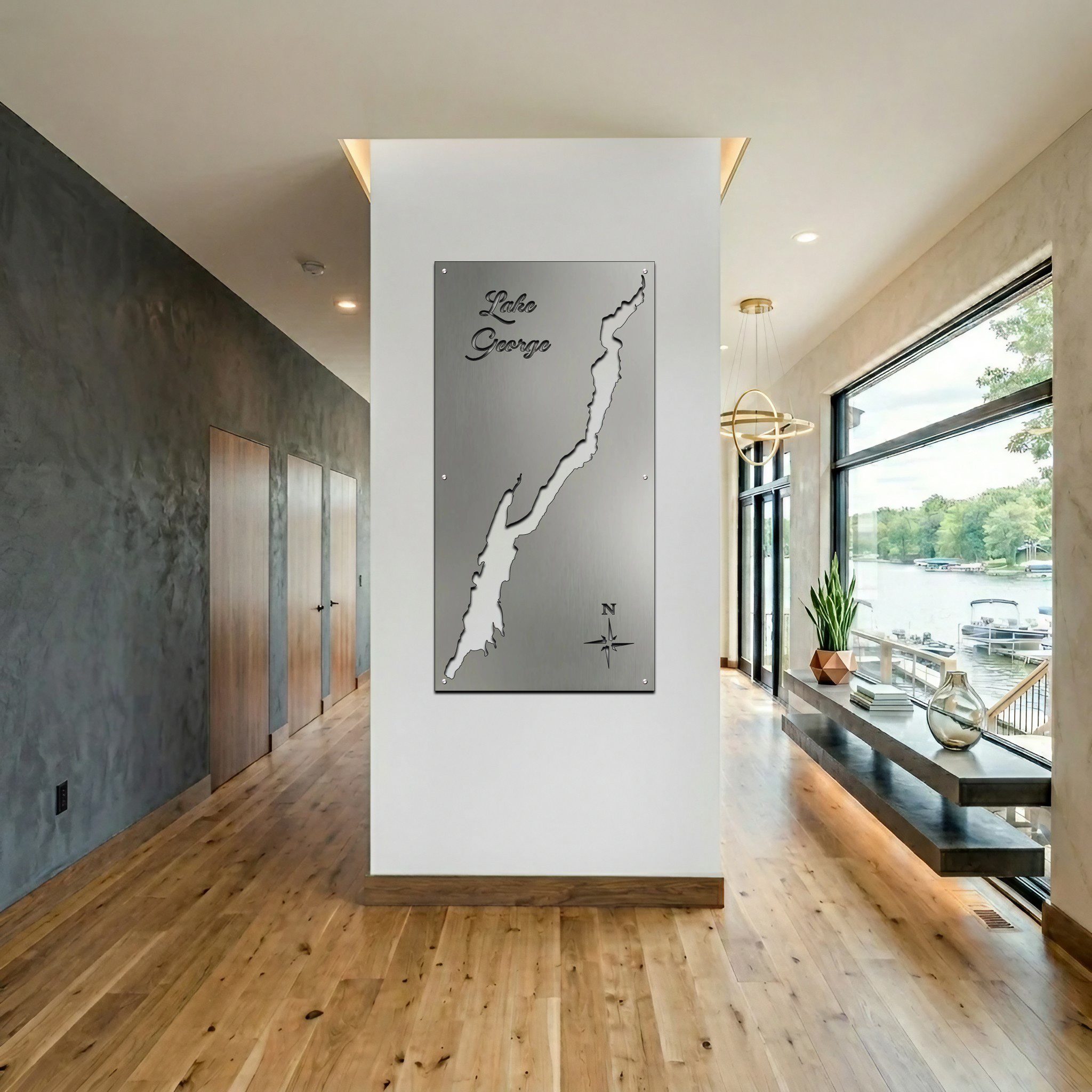 Metal map of Lake George displayed on a wall in a modern hallway. Large windows show a lakeside view.