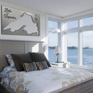 A bedroom features a bed with decorative pillows, a wall art of Lake Superior, and large windows overlooking water.